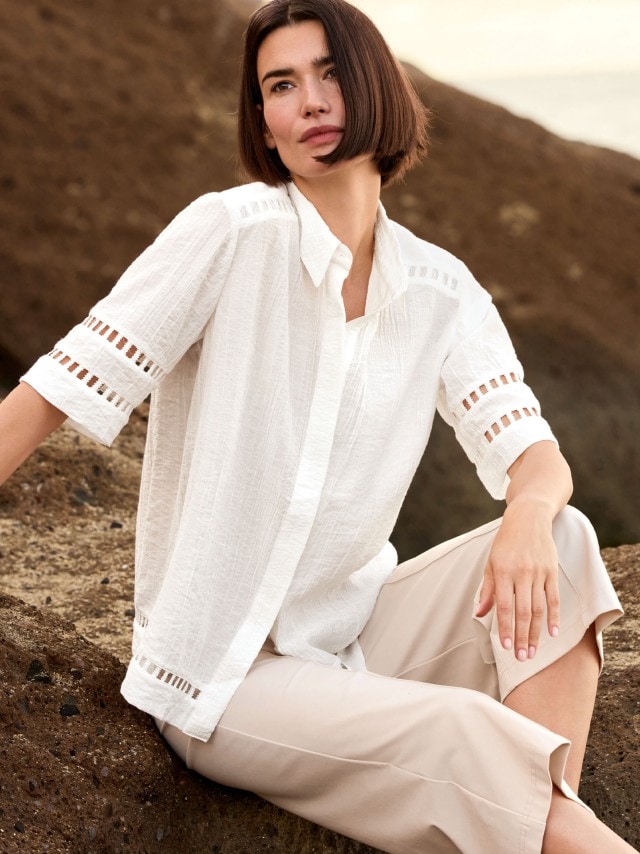 Frau sitzt auf einem Felsen an der Küste und trägt eine Statement Bluse in Weiß, dazu eine Hose in Beige