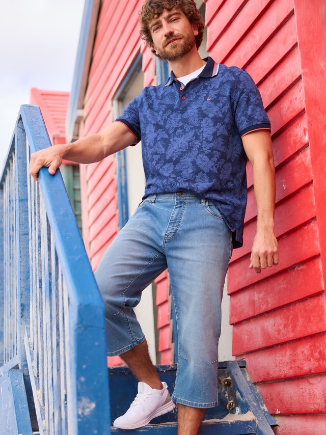 Ein Mann trägt ein dunkelblaues Poloshirt mit floralem Muster, kombiniert mit hellblauen, langen Jeansshorts und weißen Sneakern. Der Look wirkt sportlich, sommerlich und entspannt.