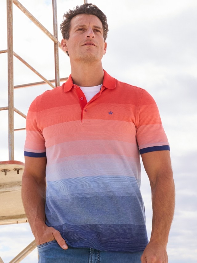 Ein Mann trägt ein sommerliches Poloshirt mit Farbverlauf von Rot über Rosa bis Blau und kombiniert es mit einer Jeans. Der Look ist frisch, modern und perfekt für entspannte Tage am Strand.