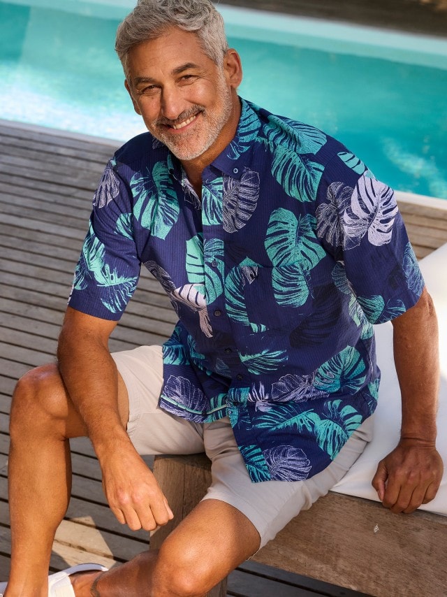 Ein Mann sitzt am Pool und trägt ein kurzärmliges, blaues Hemd mit großem, grünem und weißem Blätterdruck sowie beigefarbene Shorts.