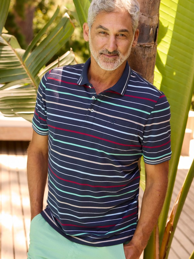 Ein Mann steht draußen vor Pflanzen und trägt ein dunkelblaues Poloshirt mit bunten Streifen sowie eine hellgrüne Hose.
