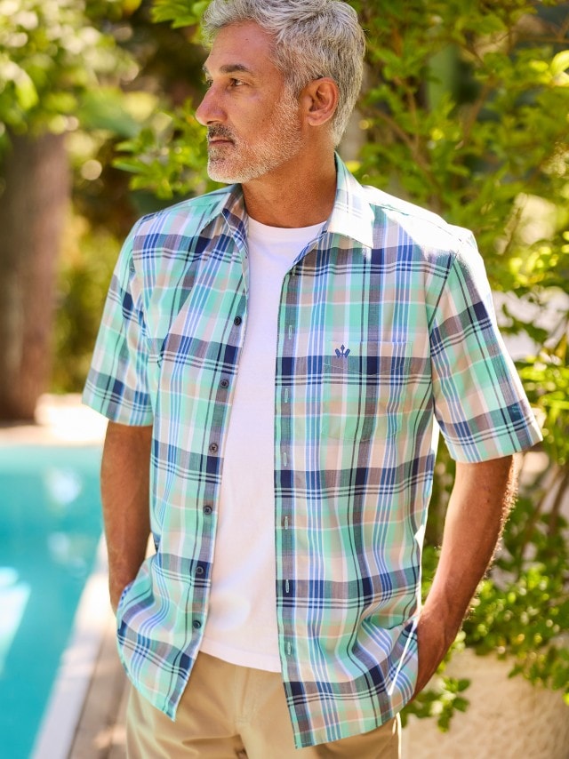 Ein Mann steht draußen am Pool und trägt ein kurzärmliges, mintgrün-blau kariertes Hemd über einem weißen T-Shirt und eine beige Hose.