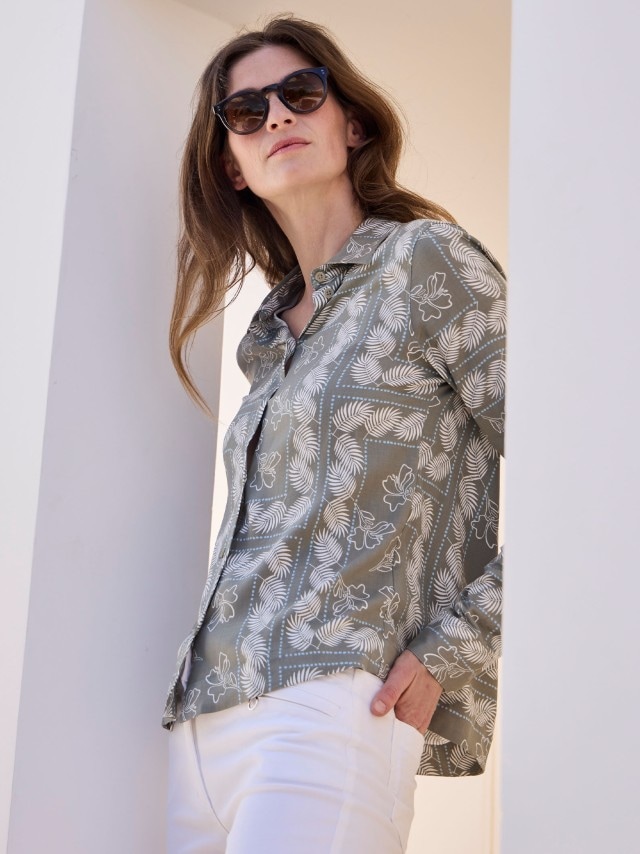 Eine Frau mit Sonnenbrille trägt eine gemusterte Bluse und eine weiße Hose.
