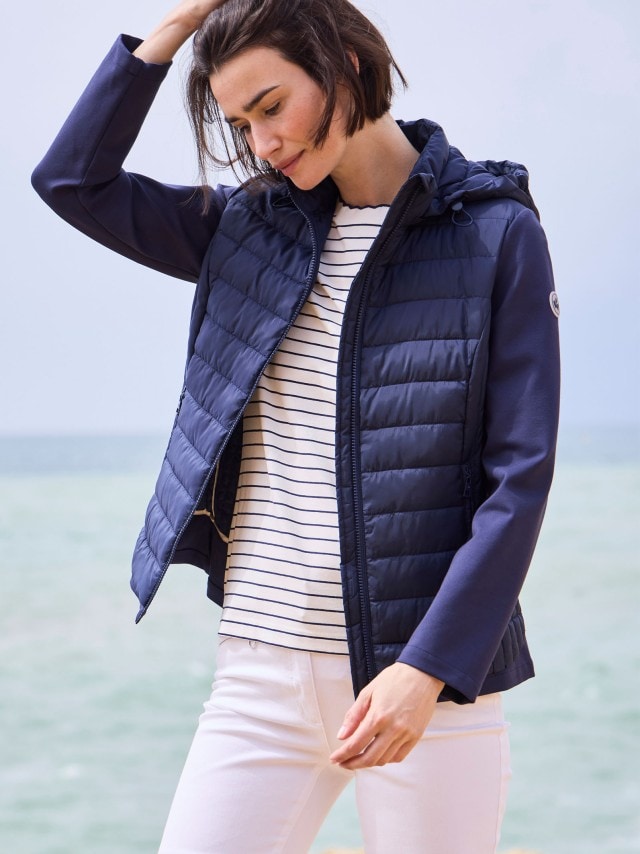 Eine Frau steht am Strand und trägt eine dunkelblaue Steppjacke, ein gestreiftes Shirt und eine weiße Hose.