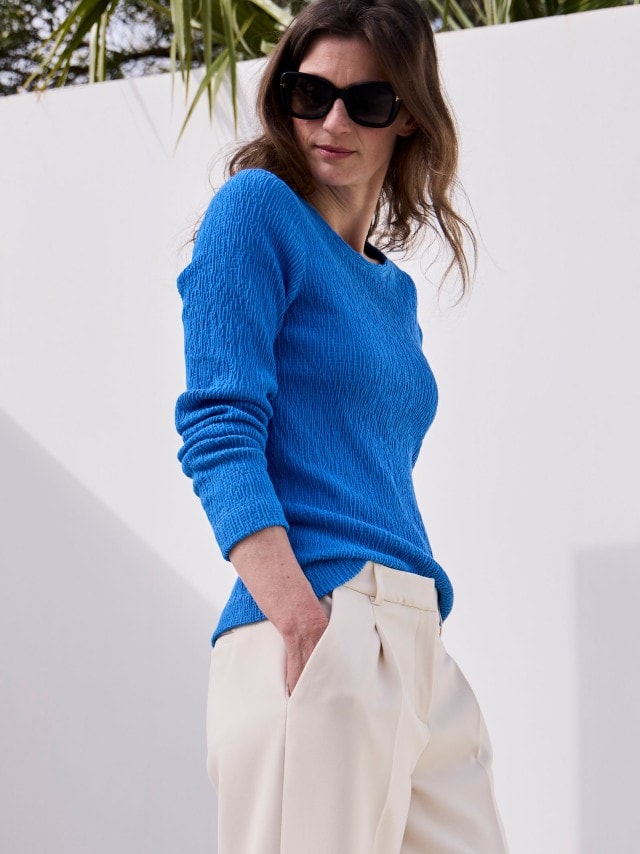 Eine Frau mit Sonnenbrille trägt einen blauen Pullover und eine helle Stoffhose.