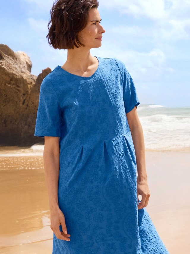 Eine Frau trägt ein blaues, kurzärmliges Kleid mit dezentem Muster und steht am Strand.