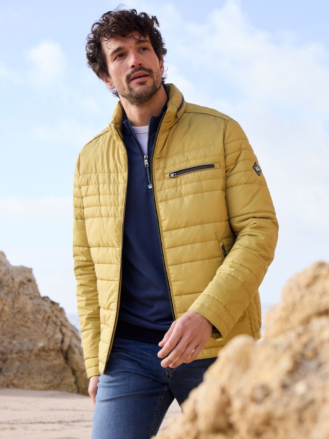 Ein Mann an der Küste trägt eine gelbe Leichtjacke, darunter einen Pullover in Marine und dazu eine blaue Jeans.