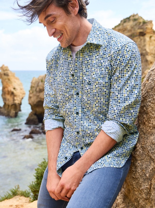 Ein Mann steht an einem Felsen am Strand und trägt ein gemustertes Hemd, dazu eine Jeans. 
