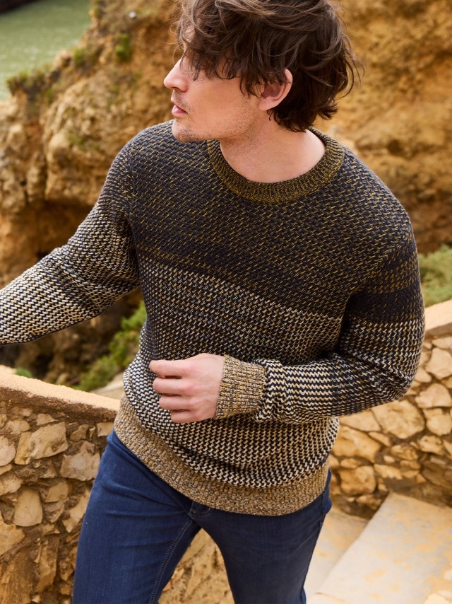 Ein Mann steht an einer Felsküste und trägt einen Strickpullover, dazu eine Jeans.