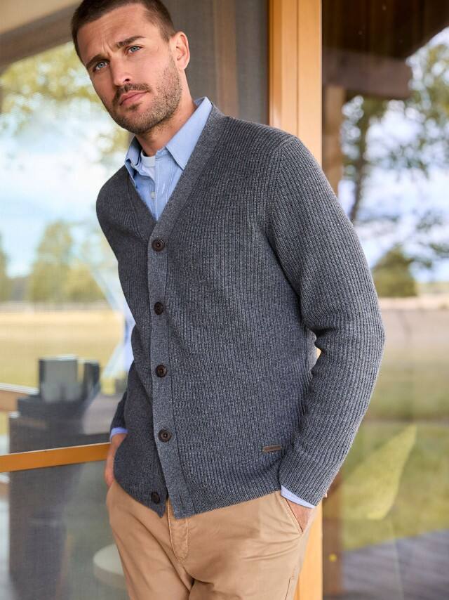 Ein Mann steht vor einer großen Fensterfront und trägt ein hellblaues Hemd, darüber einen dunklen Patentstrick-Cardigan und dazu eine Hose in Beige.