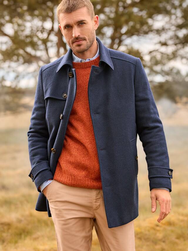 Ein Mann trägt eine Wollmix Cabanjacke in Marine, darunter einen Strickpullover in Orange und dazu eine Hose in Beige.