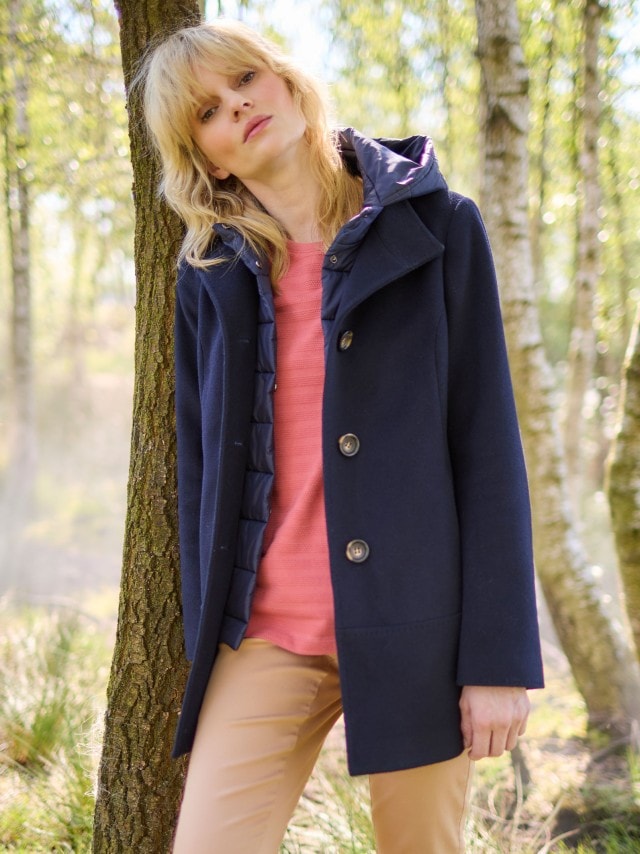 Ein Frau steht vor einem Baum und trägt die Wolljacke mit Steppeinsatz in Marine, darunter ein Baumwollshirt in Siena und dazu eine Hose in Beige.