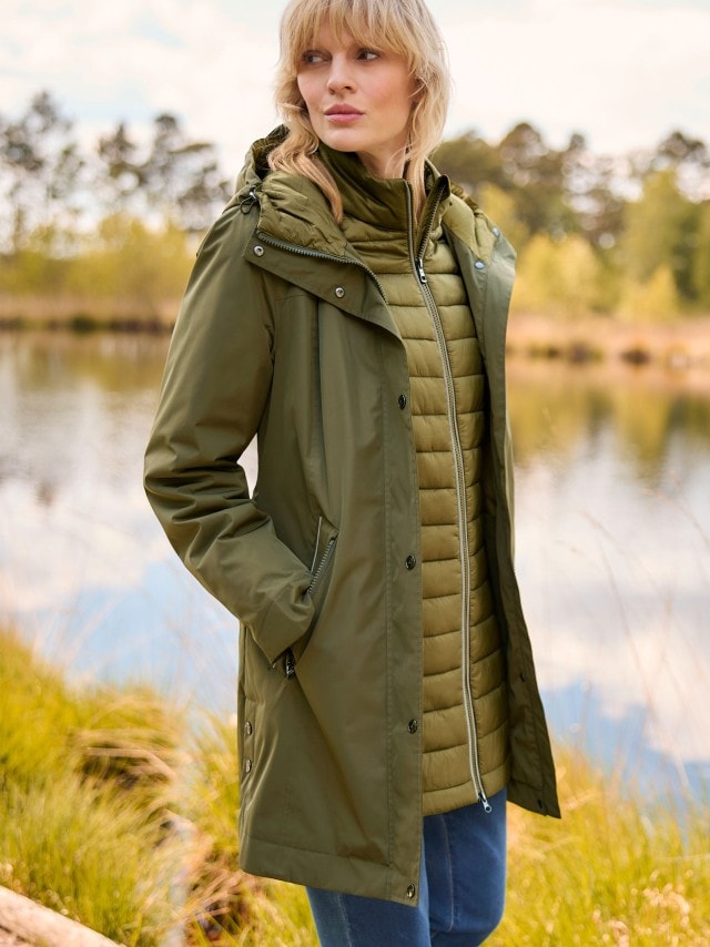 Eine Frau steht vor einem See und trägt den Aquastop Parka 3-in-1 in Oliv dazu eine Jeans.