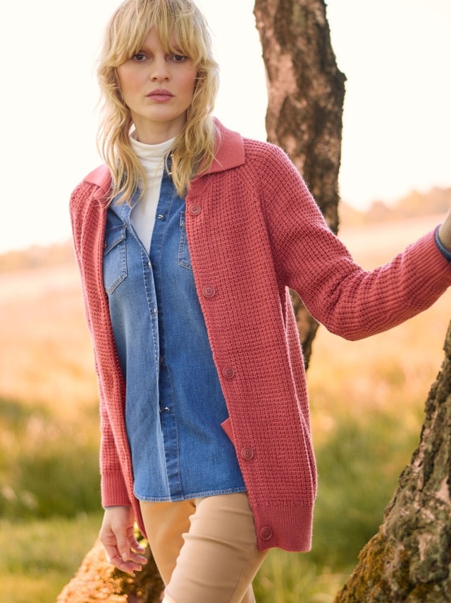 Ein Frau steht vor einem Baum und trägt die Merino-Strickjacke Waffelstruktur in Siena darunter eine Jeansbluse mit Rollkragenshirt und dazu eine Hose in Beige.