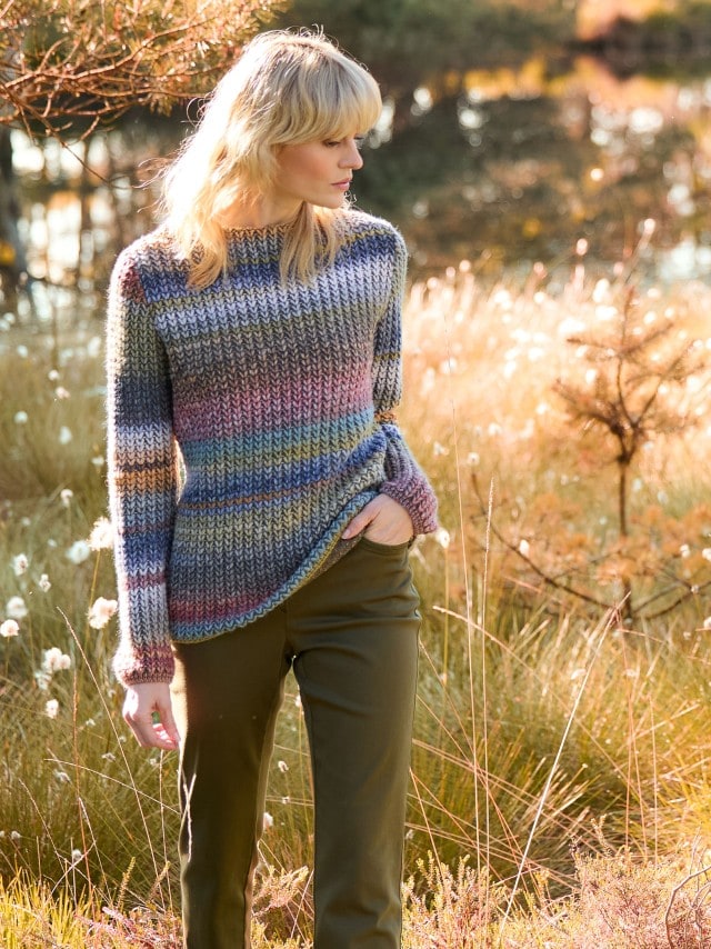 Eine Frau steht auf einer herbstlichen Wiese, schaut zur Seite und trägt einen Grobstrick-Pullover Farbverlauf, dazu eine dunkele Hose.