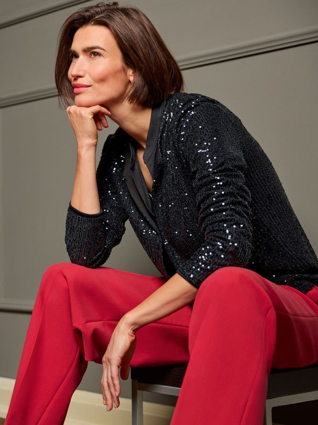 Eine Frau sitzt auf einem Stuhl, ist nach vorne gelehnt und trägt eine schwarze Paillettenjacke, dazu eine rote Hose.