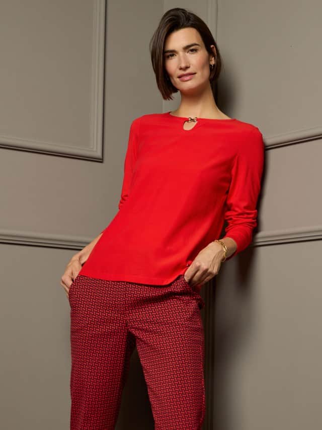 Eine Frau steht vor einer dunklen Wand und trägt ein rotes Viskoseshirt, dazu eine rotgemusterte Hose. 