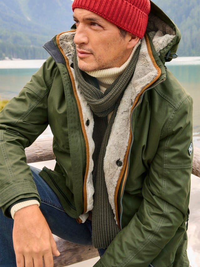 Ein Mann hockt an einem Bergsee und trägt die Aquastop Winterjacke in Oliv, darunter eine Rollkragen-Pullover mit Schal, eine Jeans und eine rote Mütze.