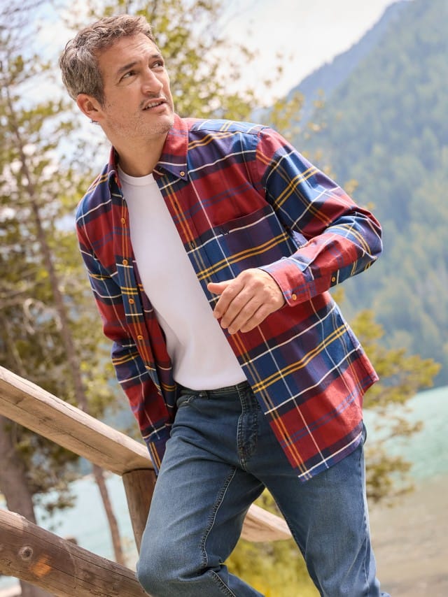 Ein Mann steht an einem Holz-Geländer und trägt ein offenes Diamanten Twill-Hemd in Karo Bordeaux, darunter ein weißes Shirt und dazu eine Jeans.