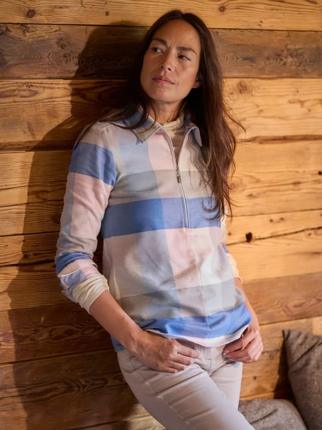 Eine Frau steht an einer Holzwand und trägt die Kuschelflanell Zip-Bluse in Karo Altrosa, dazu eine Hose in Beige.