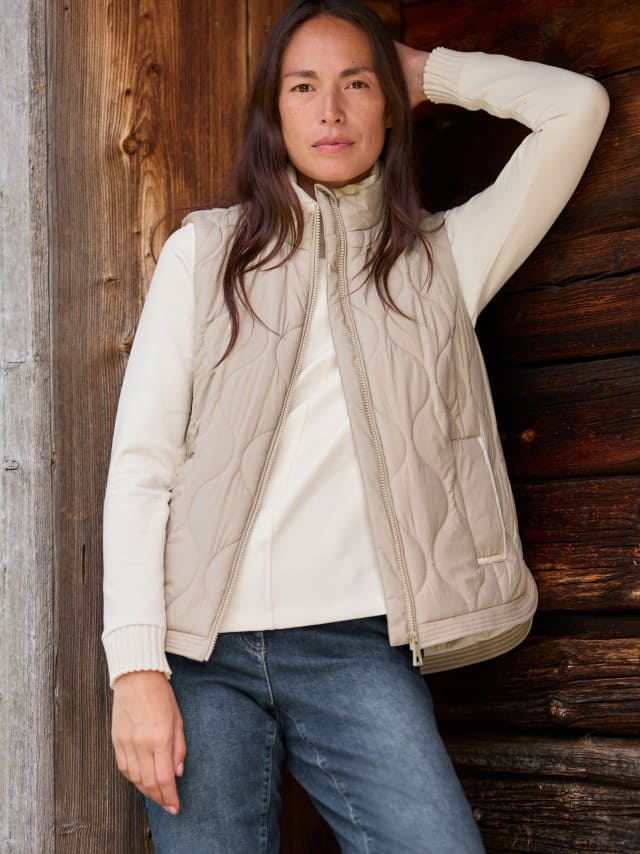 Eine Frau steht vor einer Holzwand und trägt die Wellensteppweste in Toffee/Creme, darunter ein weißes Sweatshirt und dazu eine Jeans.