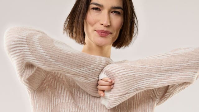 Eine Frau trägt einen kuscheligen, hellrosa-beigen Strickpullover mit breiten Bündchen und leichtem Muster für einen weichen, gemütlichen Look.
