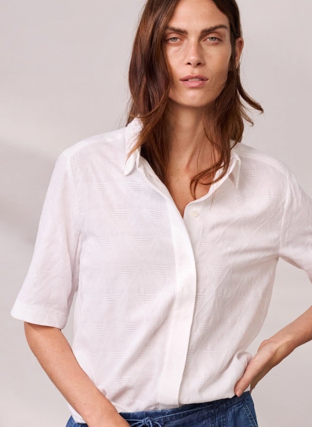 Frau trägt eine weiße, locker geschnittene Bluse mit dezentem geometrischem Muster und halblangen Ärmeln, kombiniert mit einer Jeans. Der Look wirkt frisch und modern.