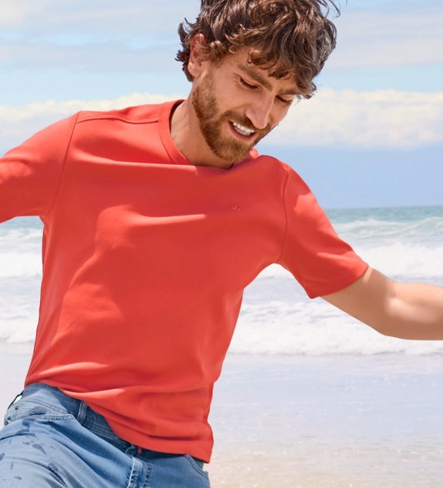Ein Mann mit lockigen Haaren trägt ein rotes T-Shirt und eine helle Jeans  und läuft lachend am Strand entlang.