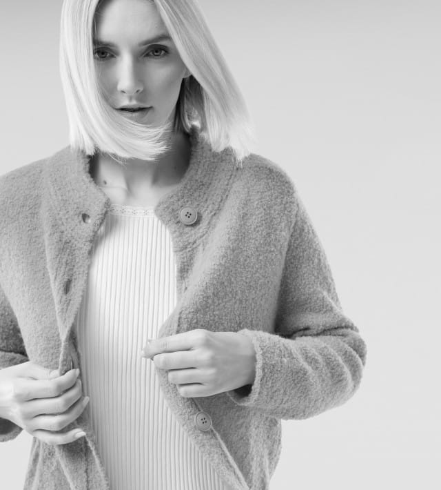 Eine blonde Frau trägt ein weißes Ripp-Shirt und darüber eine Kuschel-Strickjacke.