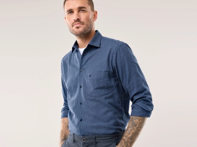 Ein Mann trägt ein Flanellhemd in blau, darunter ein weißes Shirt.