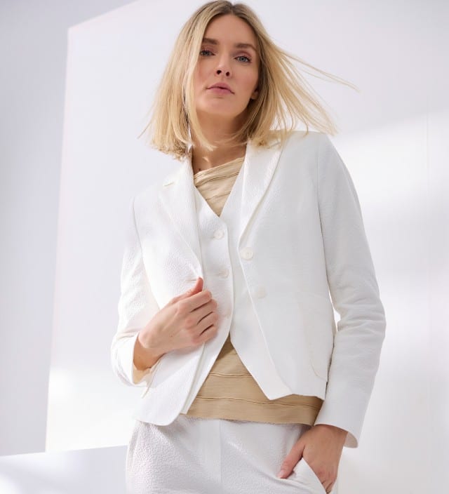 Eine blonde Frau trägt einen weißen Seersucker Blazer, mit passender Weste und Hose, dazu ein Baumwollshirt mit Streifenstruktur in Beige. 