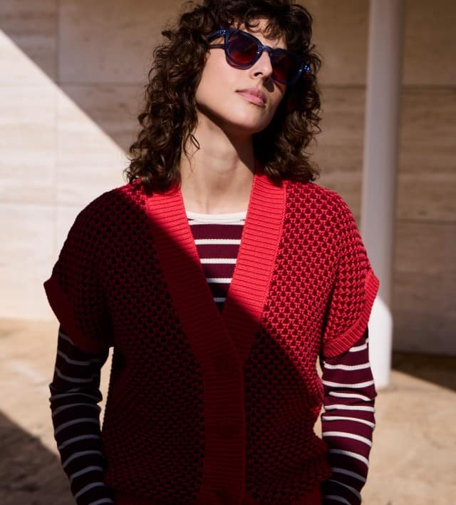 Eine Frau mit lockigem Haar und Sonnenbrille, trägt eine Strickweste mit Waffelstruktur in Signalrot, darunter ein passendes gestreiftes Shirt.