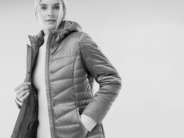 Eine Frau trägt eine gesteppte Winterjacke mit Kapuze und darunter einen grob gestrickten Pullover. Ihr Stil wirkt warm, modern und alltagstauglich.