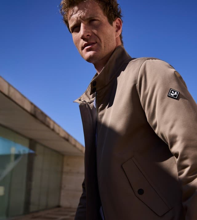 Ein Mann vor einem Natursteingebäude trägt einen graubraunen Freizeitblouson, dazu eine Jeans.