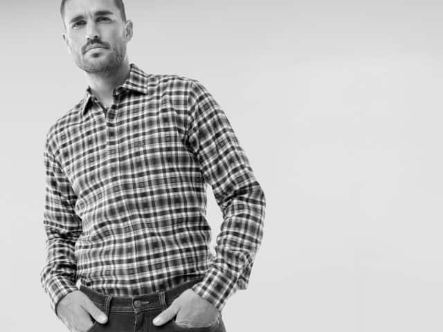 Ein Mann trägt ein kariertes Hemd mit langen Ärmeln und eine dunkle Jeans. Sein Stil wirkt lässig und zugleich gepflegt.
