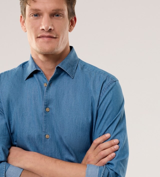 Ein Mann trägt ein hellblaues Denim-Hemd und verschränkt die Arme.
