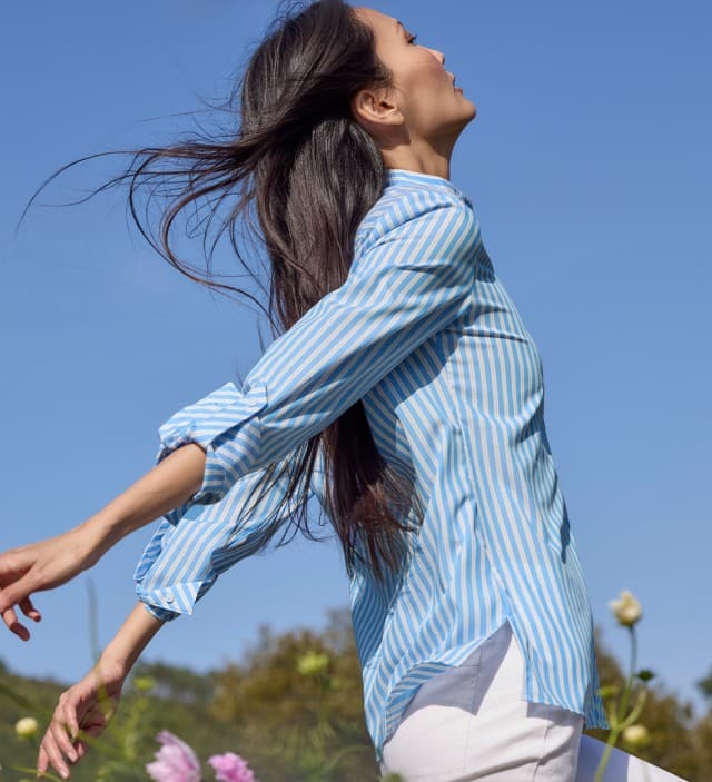 Eine Frau tanzt auf einer Blumenwiese und trägt eine gestreifte Bluse in Blau/Weiß, dazu eine weiße Hose.