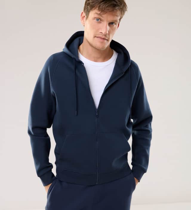 Ein Mann trägt eine Sweatshirt-Jacke mit Kapuze in Marine, dazu die passende Hose und ein weißes Shirt.