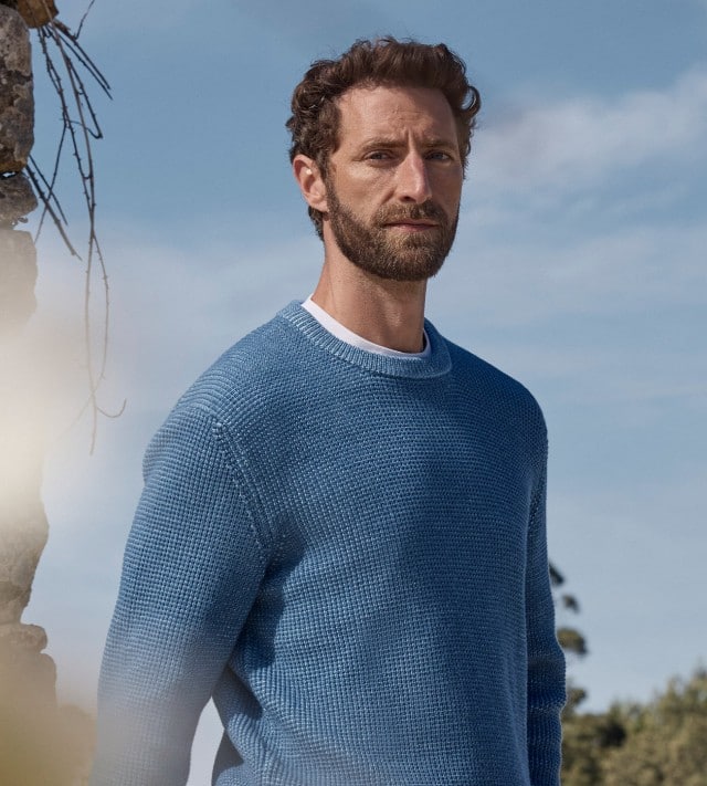 Ein Mann trägt einen Strukturpullover Bicolor in Jeansblau und eine Frau ein Baumwollshirt Ringelstruktur in Offwhite/Nachtblau, im Vordergrund sind gelbe Blüten zu sehen.