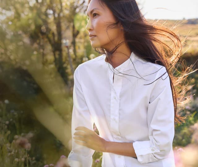 Eine Frau steht auf einer Frühlingsweide und trägt eine weiße Bluse mit Stehkragen.
