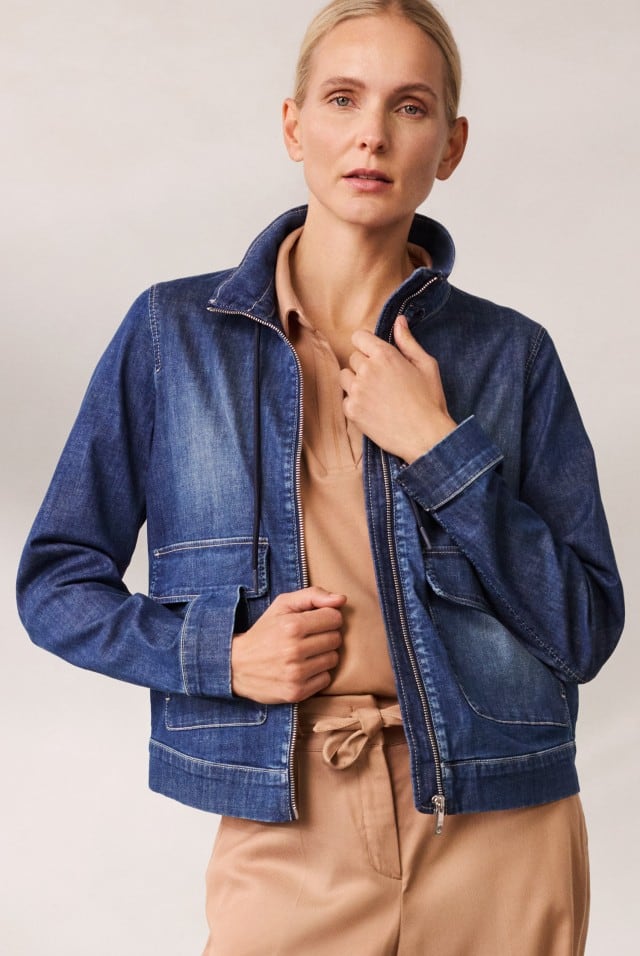 Eine Frau trägt eine blaue Jeansjacke über einem beigefarbenen Shirt und Hose.