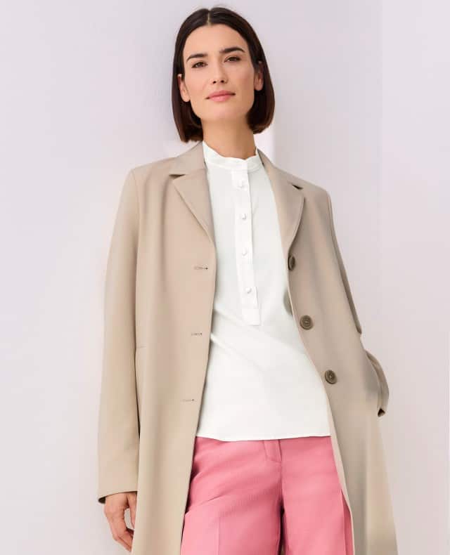 Eine Frau vor einem hellen Hintergrund, trägt eine weiße Bluse mit Stehkragen, darüber einen Koffer-Mantel in Sand und dazu eine Hose in der Farbe Rosenholz.