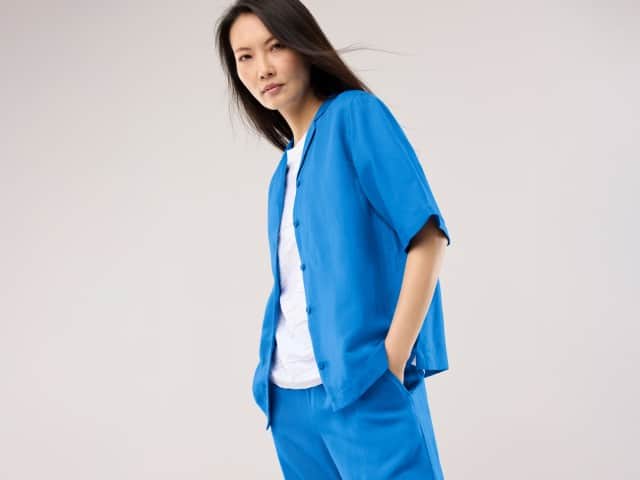 Eine Frau trägt ein blaues Leinen-Set bestehend aus Hemd und Hose, dazu ein weißes Shirt. 