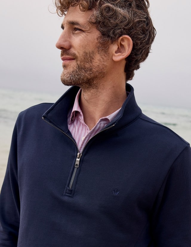 Eine Mann am Strand trägt einen dunkelblauen Troyer über einem gestreiften Hemd, was modern, entspannt und dezent sportlich wirkt.