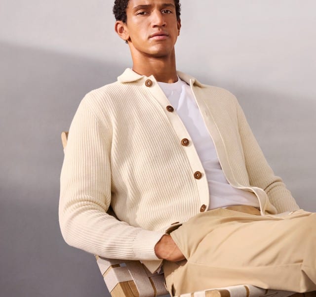 Ein Mann sitzt lässig auf einem Stuhl und trägt einen gerippten Polo-Cardigan in Wollweiß, darunter ein weißes Shirt und dazu eine passende Hose.