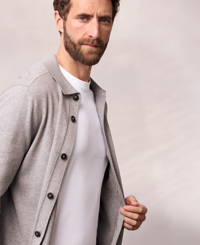 Ein Mann trägt ein geknöpftes Strickpolo in Grau, darunter ein weißes Shirt.