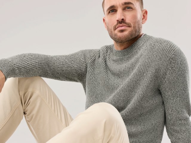 Ein Mann sitzt lässig auf dem Boden und trägt einen grauen Strickpullover, dazu eine helle Hose.