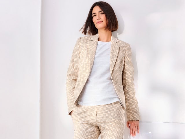Eine Frau trägt einen Jacquardblazer in der Farbe Sand, darunter ein weißes Baumwollshirt mit Streifenstruktur und dazu eine passende Hose.
