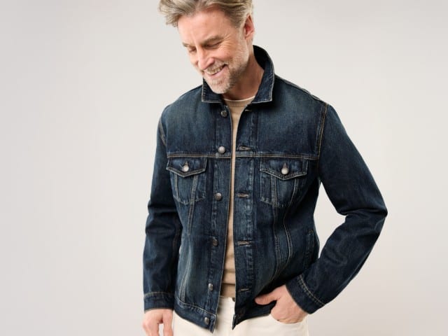 Ein Mann trägt eine Jeansjacke, darunter einen Pullover in Beige und dazu eine weiße Jeans.