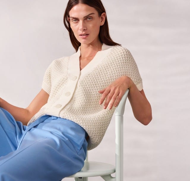 Ein Frau sitzt lässig auf einem Stuhl und trägt eine Strickweste mit Waffelstruktur in Offwhite, dazu eine blaue Hose.